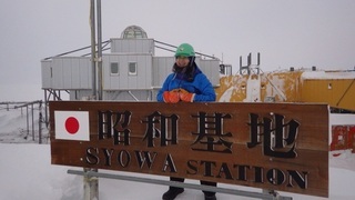 写真：昭和基地の立て看板に手をついて写真におさまる渡貫淳子氏